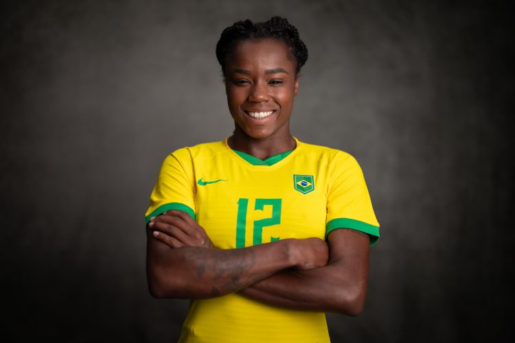 A atacante Ludmila está vestida com a camisa amarela da Seleção Brasileira e aparece sorrindo, com os braços cruzados